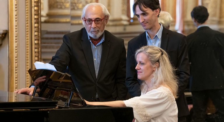 Un mujer al piano y dos hombres leyendo junto a ella la partitura